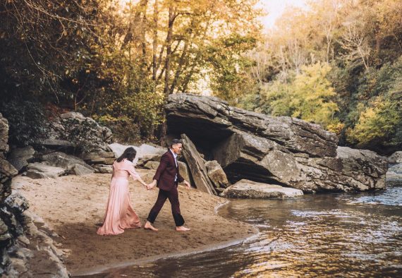 Couple dresses in formal attire walking together outdoors by a river elopement