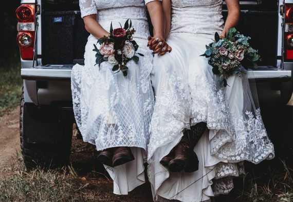 Two brides sitting in the back of a truck wearing boots and holding bouquets